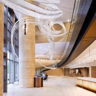 A stunning large chandelier graces a sleek hotel lobby, showcasing innovative custom lighting design. This big chandelier features graceful, ribbon-like strands of illuminated material that swoop and curl through the air, creating an ethereal effect. The flowing lights contrast beautifully with the warm-toned cylindrical pillars and expansive windows. Below, a modern reception area with a backlit counter adds to the luxurious ambiance. This unique hotel chandelier transforms the spacious lobby into a captivating blend of artistry and functionality, exemplifying how bespoke lighting can elevate hospitality spaces to new levels of sophistication and visual appeal.