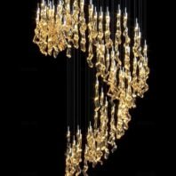 A stunning large chandelier exemplifies exquisite custom lighting design for luxury hotels. This big chandelier features a dramatic cascade of golden, organically-shaped glass elements suspended at varying lengths. The illuminated pieces create a flowing, waterfall-like effect, descending in a graceful curve against a black background. Each glass element glows warmly, resembling molten gold or crystallized honey. The intricate arrangement transforms the space with its dynamic play of light and shadow, offering a breathtaking focal point that elevates the hotel's ambiance to new heights of opulence and sophistication.