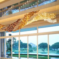 A magnificent custom lighting hotel chandelier adorns a spacious lobby with floor-to-ceiling windows overlooking a lush tropical landscape. The chandelier features a flowing, wave-like design composed of thousands of glass elements in various golden hues, transitioning from deep amber to light gold and clear. Above the main chandelier, a band of blue and white lighting creates a sky-like effect. The warm glow of the chandelier contrasts beautifully with the cool, natural scenery outside, which includes palm trees, a manicured lawn, and a glimpse of a pool. This unique lighting installation transforms the hotel space into an opulent gateway between interior luxury and exterior paradise.