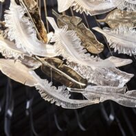 A close-up of a large custom lighting hotel chandelier featuring intricate glass feathers. This big chandelier showcases delicate, translucent feathers in clear and golden hues, suspended by thin wires. The feathers appear to float gracefully, creating a mesmerizing, ethereal effect. This unique custom lighting design adds a touch of whimsy and elegance to luxury hotel spaces, transforming them into enchanting environments.