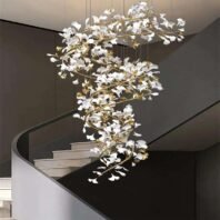 A stunning spiral chandelier featuring white glass petals and gold-toned branches, suspended in a modern stairwell. The chandelier has an organic, tree-like structure that twists downward in a graceful spiral. Numerous white glass elements, resembling stylized flowers or leaves, are attached to the golden metallic framework. The fixture creates a dramatic cascade effect, filling the vertical space of the stairwell. The chandelier's warm, golden glow contrasts beautifully with the dark gray walls of the stairwell. A portion of a curved, modern staircase with light-colored steps is visible in the lower right corner of the image. The chandelier's intricate design and the interplay between the white glass and gold metal create a luxurious and eye-catching focal point in this contemporary interior space.