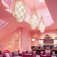 A luxurious hotel lounge featuring custom lighting with large chandeliers resembling netted eggs. These big chandeliers, three in total, have white oval shades wrapped in golden chain-like netting, suspended from the ceiling. The custom lighting perfectly complements the room's pink color scheme, including plush velvet sofas, a curved staircase, and paneled walls adorned with framed artwork. The space exudes elegance with its bar area, coffee table books, and skylights, creating a sophisticated atmosphere enhanced by the unique chandelier design.