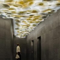 A striking custom lighting installation featuring a large chandelier that spans the entire ceiling of an elevator lobby. This hotel chandelier consists of numerous organic, cloud-like shapes made of translucent material, each softly illuminated from within. The big chandelier creates a mesmerizing, undulating pattern of light and shadow, resembling a luminous sky. Warm golden hues emanate from the center of each cloud-like form, contrasting beautifully with the dark, textured walls. A person standing below, gazing upward, emphasizes the grand scale and captivating nature of this unique lighting design.