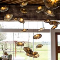 A unique custom lighting installation featuring multiple glass elements suspended from the ceiling. Each piece resembles an organic, petal-like form made of translucent glass with subtle smoky tones. The glass elements vary in size and shape, creating a dynamic, floating composition. Warm light emanates from within each piece, giving them a golden glow that contrasts beautifully with their translucent nature. The installation hangs from a rustic wooden beam ceiling, adding a touch of modern elegance to the natural, earthy backdrop. Large windows in the background offer a view of a serene outdoor landscape, blurring the line between interior and exterior spaces. This innovative lighting design transforms the room into an artistic display, evoking the feeling of being under a canopy of delicate, illuminated leaves or flower petals. The overall effect is both ethereal and grounding, perfect for a space that aims to blend natural elements with contemporary design.