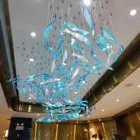 A spectacular large hotel chandelier showcasing innovative custom lighting design. This big chandelier features hundreds of blue and clear glass fish-shaped elements suspended from the ceiling. The fish appear to be swimming in a school, creating a dynamic, flowing pattern across the entire ceiling. Each glass piece catches and reflects light, mimicking the shimmer of fish scales underwater. This stunning custom lighting installation transforms the hotel space into an underwater wonderland, perfect for luxury establishments seeking a unique, immersive centerpiece that captivates guests with its oceanic beauty.