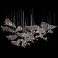 A striking large hotel chandelier showcasing innovative custom lighting design. This big chandelier features numerous clear glass elements shaped like abstract leaves or petals, suspended at varying heights by thin wires against a black background. The glass pieces appear to float in mid-air, creating a dynamic, organic composition reminiscent of a flock of birds or a school of fish in motion. Small light sources are interspersed among the glass elements, casting a soft glow and creating gentle reflections on the curved surfaces. This avant-garde piece exemplifies high-end custom lighting, perfect for creating a captivating focal point in modern, luxurious hotel spaces. The chandelier's fluid design transforms the ambiance with its light, airy presence, blending artistry with functionality to create a mesmerizing sculptural lighting installation.