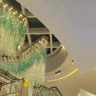 A stunning large-scale custom lighting installation in a luxury hotel atrium, viewed from below. The chandelier features multiple cascading sections of crystal-like elements in shades of emerald green and clear, creating a mesmerizing waterfall effect. The installation spans across different levels of the atrium, with each section suspended at varying heights from the curved ceiling. The crystal strands shimmer and sparkle, transitioning from vibrant green at the top to clear at the bottom, resembling a lush, ethereal forest canopy. The atrium's modern architecture is highlighted by the intricate ceiling design, featuring geometric patterns and recessed lighting. A glimpse of the structure's upper levels can be seen through the glass panels, adding depth to the space. This dramatic and luxurious lighting display transforms the hotel interior into an enchanting, nature-inspired environment.