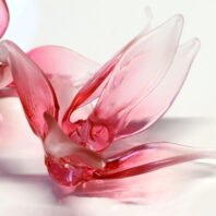 A close-up of a stunning pink glass element, likely part of a large custom chandelier for high-end hotel spaces. This intricate piece resembles a blooming flower or an abstract bird, with multiple translucent petals or wings in various shades of pink. The glass has a fluid, organic form with graceful curves and delicate edges. Light plays beautifully across the surface, creating a gradient from deep rose to pale blush. This bespoke lighting component showcases the artistry in custom chandelier design, perfect for adding a touch of elegance and warmth to luxury interiors.