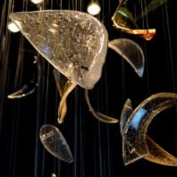 A close-up of a large custom chandelier, ideal for luxury hotel interiors. This big chandelier features an array of suspended glass elements in various shapes and sizes. The foreground showcases a large, curved glass piece with intricate bubble-like inclusions, creating a mesmerizing texture. Other glass forms, including golden-hued and clear crescent shapes, hang from thin wires, adding depth and complexity to the design. The interplay of light on these custom lighting elements creates a captivating, ethereal atmosphere, perfect for enhancing the ambiance of grand spaces.