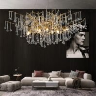 A contemporary living room featuring a stunning gold branch crystal chandelier as the centerpiece. The chandelier has an organic, tree-like structure with golden branches adorned with numerous crystal droplets, creating a sparkling effect. It hangs above a large, L-shaped beige sectional sofa decorated with various throw pillows in muted tones. A white rectangular coffee table sits in front of the sofa, and a plush gray ottoman is visible to the side. The room has a dark, textured wall with vertical ribbing, against which hangs a black and white portrait photograph. The floor features a patterned area rug in gray tones. The overall design combines modern minimalism with luxurious elements, creating a sophisticated and elegant living space.