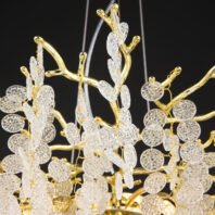 Close-up of an exquisite chandelier featuring gold-plated branches adorned with textured crystal discs. The image shows the intricate design of the fixture, with polished gold-toned metal forming organic, tree-like branches against a dark background. Numerous clear crystal elements hang from these branches, each with a unique, dimpled texture resembling water droplets or ice crystals. The crystal discs are connected to the golden structure by thin, golden wires, creating a delicate and airy appearance. Thin suspension wires are visible at the top of the image. The combination of the reflective gold surface and the translucent, light-catching crystal elements creates a luxurious and sophisticated aesthetic. The chandelier's design mimics the organic forms of nature, resulting in a harmonious blend of elegance and natural beauty.