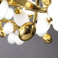 Detailed view of a contemporary chandelier featuring frosted white glass orbs and polished gold elements. The image focuses on a highly reflective, egg-shaped golden ornament hanging from a sleek gold-plated arm. In the background, several frosted white glass spheres are visible, creating a bubble-like effect. The gold-toned metal structure of the chandelier is prominently displayed, showcasing its smooth, modern design. The contrast between the matte white glass globes and the shiny gold surfaces creates a luxurious and sophisticated aesthetic. The chandelier's design combines organic, rounded shapes with precise metalwork, resulting in a fixture that balances warmth and elegance with contemporary style.