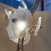 Close-up of a single glass flower element from a chandelier. The glass shade is large, circular, and has a rippled or textured surface resembling a sea anemone or a stylized flower petal. It's translucent with a frosted appearance, allowing light to diffuse through it. At the center of the glass shade is a small, dark, spherical ornament. The glass flower is attached to a golden branch or stem, which appears to be part of a larger, tree-like structure. In the background, parts of other similar glass flowers are visible, suggesting this is one piece of a larger, more elaborate chandelier. The image captures the intricate details of the glass work and the organic, nature-inspired design of the lighting fixture.