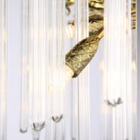 Close-up of a luxurious chandelier featuring elongated glass tubes and an ornate gold light fixture. The image showcases several clear, cylindrical glass pendants hanging vertically, with ribbed or striated textures giving them a delicate, translucent appearance. In the center, a prominent gold-plated fixture with an intricate diamond-patterned design is visible, housing a bright light bulb. The gold element resembles a stylized leaf or bud shape. The glass tubes refract light, creating a soft, ethereal glow against the light background. This chandelier combines sleek, contemporary design in the glass elements with more traditional, ornate detailing in the gold fixture, resulting in a striking piece that bridges modern and classic aesthetics.