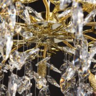 Close-up view of a luxurious chandelier's central structure, showcasing golden branches and crystal pendants. The image reveals the intricate design of the chandelier's core, with a gold-plated central disc from which ornate golden branches radiate outward. Crystal pendants of various shapes and sizes hang from thin wires, creating a shimmering cascade effect. The crystals refract light, producing rainbow-like prisms and sparkles. Gold-toned decorative elements, including spiral and leaf-like forms, are interspersed among the crystal drops. This detailed perspective highlights the chandelier's exquisite craftsmanship, blending organic shapes with opulent materials. The interplay of light on the gold and crystal elements creates a dazzling display, emphasizing the fixture's role as both a functional light source and a piece of sculptural art.