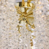 A close-up image of the central structure of a luxurious chandelier. The main body is made of polished gold-toned metal, featuring a smooth, cylindrical top section. Extending from this are intricately designed branches that mimic organic, tree-like forms. These golden branches spread outward and downward, creating a natural, flowing appearance. Suspended from the gold elements are numerous strands of clear crystals, forming cascading columns that catch and reflect light brilliantly. The crystals appear to be small, faceted pieces, creating a shimmering, icicle-like effect. Warm light is visible, emanating from within the structure and illuminating the crystals, causing them to sparkle against the background. The combination of the sleek, reflective gold and the sparkling crystals results in a striking contrast of textures and materials, embodying both natural inspiration and opulent design.