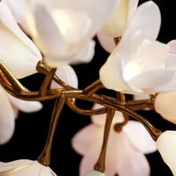 Close-up of an elegant branch chandelier featuring delicate white and pale pink flower-shaped lights. Golden metallic branches intertwine gracefully, supporting the blooming fixtures. The chandelier's intricate design mimics natural tree branches, creating a stunning blend of nature-inspired artistry and luxurious lighting. Set against a dark background, this branch chandelier showcases exquisite craftsmanship and brings a touch of organic elegance to interior spaces.