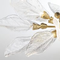 Close-up of a chandelier featuring delicate glass leaves with intricate vein patterns. The transparent leaves are attached to gold-plated fixtures, creating a nature-inspired design. Each leaf is uniquely textured, mimicking real foliage. Small light bulbs are visible within the gold fixtures, suggesting a soft, warm glow when lit. The image showcases the chandelier's blend of organic forms and luxurious craftsmanship against a light background.