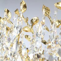 Detailed view of a chandelier featuring gold-plated leaf designs intertwined with large, faceted crystal pendants. The gold leaves have a textured, realistic appearance, while the crystals are elongated and teardrop-shaped. Light refracts through the crystals, creating prismatic effects. The intricate combination of metallic and transparent elements showcases luxury and craftsmanship in lighting design.