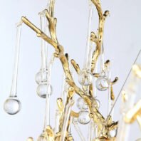 A detailed close-up view of an elegant chandelier featuring gold-plated branches interspersed with crystal droplets. The gold branches have a naturalistic, organic form resembling tree twigs or coral, with intricate textures and curves. Suspended from these golden branches are clear crystal pendants of varying lengths, each ending in a spherical crystal orb. The crystal elements are attached to the gold structure by thin, almost invisible wires, giving the impression that they're floating. The interplay between the warm gold tones and the clear, reflective crystals creates a luxurious and delicate aesthetic. This chandelier design combines nature-inspired forms with high-end materials, resulting in a piece that's both artistic and opulent.