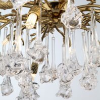 A close-up view of an opulent chandelier featuring a combination of gold-plated metalwork and intricate glass crystal elements. The upper portion of the image shows the chandelier's central structure, made of polished gold-toned metal with ornate, curved arms extending outward. From these arms hang numerous clear glass pendants, each with a unique, organic shape reminiscent of flowers or droplets. These glass elements have a delicate, hand-blown appearance with swirling patterns and textures that catch and refract light beautifully. Interspersed among the glass pendants are gold-plated accents with a textured, pineapple-like design, adding visual interest and luxury to the piece. Several small LED bulbs are visible, emitting a warm glow that illuminates the crystal elements. The interplay between the warm gold tones and the clear, sparkling glass creates a stunning visual effect. This chandelier combines classical elegance with modern design elements, resulting in a piece that would serve as a captivating focal point in any high-end interior space.