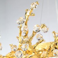 A close-up view of an exquisite chandelier detail featuring gold-plated branches adorned with crystal accents. The image showcases intricate golden branches that twist and curve organically, reminiscent of tree limbs or coral formations. Each branch is meticulously crafted with a smooth, polished finish that gleams brightly. Attached to these golden branches are numerous faceted crystal orbs of varying sizes, resembling sparkling dewdrops or blossoms. These crystals are precisely cut, allowing them to catch and refract light brilliantly, creating a dazzling display of rainbow-like reflections. The crystals are strategically placed at the ends of branches and at junction points, enhancing the organic, floral-inspired design. In the background, a portion of what appears to be a larger gold-textured surface is visible, suggesting this detail is part of a more extensive, ornate chandelier structure. The combination of the warm, lustrous gold with the clear, sparkling crystals creates a luxurious and elegant aesthetic, perfect for high-end interior spaces. This chandelier element beautifully merges nature-inspired design with opulent materials, resulting in a piece that is both a functional light fixture and a stunning work of art.