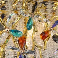 A close-up, detailed view of an opulent chandelier featuring a harmonious blend of gold and multi-colored crystal elements. The structure is composed of golden branches that curve and intertwine organically, reminiscent of tree limbs or vines. Attached to these branches are flower-like formations made from faceted crystals in various vibrant colors including turquoise, amber, clear, and deep blue. Each crystal flower has petals of different colors and shapes, creating a dynamic, jewel-like effect. The centers of these crystal flowers are adorned with small gold spheres, adding to their floral appearance. Surrounding and hanging from the gold branches are numerous strands of small, clear crystal droplets arranged in vertical lines, resembling a shimmering curtain of rain or icicles. The gold branches provide a warm contrast to the cool, sparkling crystals. Light reflecting off the various crystal elements creates a dazzling, multi-colored sparkle throughout the fixture. This chandelier masterfully combines nature-inspired design with luxurious materials, resulting in a piece that is both a functional light source and a stunning work of art, capable of adding drama and sophistication to any high-end interior space.