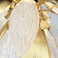 A close-up view of an exquisite chandelier featuring a mesmerizing blend of crystal and gold elements. The focal point is a large, leaf-shaped crystal piece with an intricate, ribbed texture that creates a pattern reminiscent of delicate leaf veins. This crystal element is transparent and catches light beautifully, creating a subtle sparkling effect. Surrounding the crystal leaf are gold-toned metal parts with a smooth, ribbed surface, adding a warm contrast to the cool crystal. In the background, glimpses of other crystal and gold components can be seen, including what appears to be small, bell-shaped golden casings that likely house LED lights. The image captures the chandelier's meticulous craftsmanship, showcasing the seamless integration of transparent, textured crystal with polished gold elements. This detailed view emphasizes the chandelier's nature-inspired design, combining organic forms with luxurious materials to create a stunning piece of functional art for high-end interior spaces.