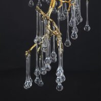 A close-up view of an exquisite branch chandelier featuring a golden, organically-shaped structure adorned with crystal pendants. The chandelier's frame is crafted from intricately designed gold-toned metal, mimicking the delicate, twisting forms of tree branches. Suspended from these golden branches are numerous clear crystal pendants of varying lengths. Each pendant consists of a long, slender stem ending in a bulbous teardrop shape, reminiscent of raindrops or icicles. The crystal elements are perfectly transparent, catching and refracting light to create a dazzling display against the dark background. This branch chandelier masterfully combines the organic beauty of nature with luxurious materials, offering a stunning focal point that bridges the gap between natural inspiration and high-end interior design. The interplay between the warm, gleaming gold branches and the cool, sparkling crystal droplets creates a mesmerizing visual effect.