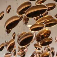 A mesmerizing custom lighting installation featuring numerous suspended copper-colored discs. These oval-shaped elements, varying in size, are arranged in a seemingly random yet harmonious pattern. Each disc has a highly polished, reflective surface that captures and bounces light, creating a warm, golden glow. The discs appear to float in space, suspended by thin, barely visible wires. This unique lighting design transforms the area into an artistic display of light and form, ideal for upscale hotel lobbies, high-end restaurants, or luxurious event spaces. The installation demonstrates how innovative custom lighting can serve as both functional illumination and a captivating focal point in sophisticated interiors.