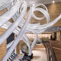 A breathtaking large chandelier dominates a luxurious hotel lobby. This big chandelier features flowing, ribbon-like strands of illuminated white material that twist and curve through the air. The custom lighting installation creates a dynamic, sculptural effect against warm wooden walls and ceilings. Below, a modern seating area with gray chairs sits on a patterned carpet, while a spiral staircase adds architectural interest. This innovative hotel chandelier transforms the space into an awe-inspiring blend of artistry and functionality, showcasing the impact of bespoke lighting design in creating unforgettable hospitality experiences.