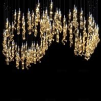 A spectacular large chandelier showcases innovative custom lighting design for luxury hotels. This big chandelier features hundreds of golden, organically-shaped glass elements suspended at varying lengths, creating an undulating wave pattern. Each piece is illuminated, resembling a shower of golden raindrops or a constellation of stars. Thin wires and small LED lights add depth and sparkle. Set against a black background, this mesmerizing hotel chandelier transforms the space into a celestial wonderland, offering a breathtaking focal point of warmth and opulence.