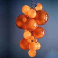 A striking custom lighting installation featuring a large hotel chandelier composed of multiple glass spheres in various shades of orange. This big chandelier showcases an array of glossy, translucent orbs clustered together, ranging from small to large sizes. The custom lighting piece is suspended by a chain, creating a playful, bubble-like arrangement against a contrasting dark blue background. The warm orange hues of the glass spheres emit a soft, inviting glow, perfect for adding a pop of color and modern flair to contemporary spaces.