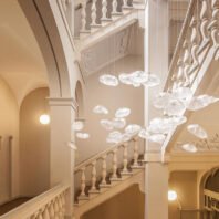 A breathtaking custom lighting installation graces a grand classical stairwell. Multiple translucent, cloud-like forms are suspended from thin wires, creating the illusion of floating illuminated clouds. These organic shapes vary in size and are clustered at different heights, filling the void of the stairwell with a dreamy, ethereal presence. The soft, diffused glow of the cloud forms contrasts beautifully with the crisp white architectural elements of the space, including ornate balustrades, arched doorways, and decorative moldings. The warm, creamy tones of the walls add depth and warmth to the scene. Spherical wall sconces provide additional ambient lighting, complementing the cloud installation. This innovative lighting design transforms the traditional architectural space into a magical, contemporary art piece, seamlessly blending classical elegance with modern whimsy.