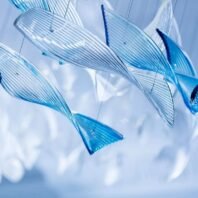 A close-up of a mesmerizing large hotel chandelier featuring custom lighting design. This big chandelier showcases multiple translucent blue glass elements shaped like elongated leaves or fins. Each piece is delicately ribbed, creating a play of light and shadow. The glass elements vary in shades of blue, from pale azure to deep sapphire, suspended at different angles to mimic the fluid motion of underwater flora or marine life. This innovative custom lighting installation transforms the space with its aquatic-inspired beauty, perfect for luxury hotels seeking a unique, calming centerpiece.