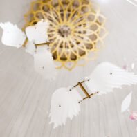 A close-up view of a large custom chandelier, perfect for luxury hotel spaces. The big chandelier features a central golden floral motif with intricate petal-like patterns, reminiscent of a sunburst design. Suspended from thin wires are delicate white glass elements shaped like butterflies, their wings spread in flight. These glass butterflies appear to float around the golden centerpiece, creating a whimsical and ethereal effect. The combination of the metallic gold flower and the translucent white butterflies showcases the artistry in custom lighting design, offering a unique and captivating visual experience for upscale interiors.