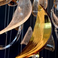 A close-up of a large custom chandelier, perfect for hotel lobbies. The big chandelier features an array of suspended glass elements in various shapes. Crescent-shaped clear glass pieces and a striking golden leaf-like form dominate the foreground. The illuminated glass creates a mesmerizing play of light, showcasing the intricate design of this bespoke lighting installation. Thin wires support the delicate glass components, adding to the chandelier's ethereal appearance and demonstrating the artistry in custom lighting solutions for luxurious spaces.