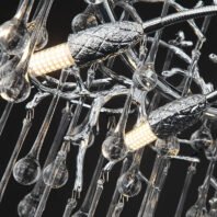 A detailed view of a contemporary chandelier featuring a complex arrangement of clear glass elements and metallic accents. The structure includes numerous thin glass rods or tubes of varying lengths, some terminating in small glass spheres. Interspersed among these are metallic elements with a textured, leaf-like appearance, possibly made of chrome or polished steel. Two cylindrical LED light sources are visible, emitting a warm glow through a honeycomb-like diffuser. The overall design is organic and fluid, reminiscent of falling water or icicles. The intricate interplay of transparent glass, reflective metal, and soft light creates a striking and modern lighting fixture against a dark background, showcasing a blend of natural inspiration and cutting-edge design.