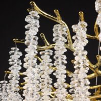 Detailed view of a luxurious chandelier featuring gold-plated branches adorned with cascading crystal elements. The image showcases the intricate design of the fixture, with polished gold-toned metal forming organic, tree-like branches. Hanging from these branches are numerous clear crystal elements, each with a unique, faceted shape that resembles cut gemstones. The crystals are arranged in vertical strings, creating a waterfall effect. The combination of the reflective gold surface and the light-catching crystal elements creates a dazzling interplay of light and shadow. The chandelier is captured against a dark background, emphasizing the contrast between the shimmering materials and the organic forms of the design.