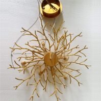 Close-up view of a chandelier's golden branch-like structure. The image shows an intricate network of gold-toned metal branches radiating outward from a central circular base. The branches are designed to mimic the organic, twisting form of tree branches or coral, creating a nature-inspired aesthetic. At the top of the image, a gold circular mounting plate is visible, from which thin wires extend to support the chandelier. The entire structure is set against a textured white background, likely a ceiling. This appears to be the base structure of a chandelier before the addition of any crystal or glass elements, showcasing the artistry and craftsmanship of the metalwork design.