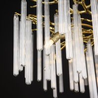 Detailed view of a luxurious chandelier featuring elongated glass tubes and golden accents. The image showcases numerous clear, cylindrical glass pendants of varying lengths hanging vertically. These glass elements have a ribbed or striated texture, giving them a delicate, translucent appearance. Interspersed among the glass tubes are golden branches with an organic, tree-like structure. A prominent gold-plated fixture with an intricate pattern is visible, likely part of the chandelier's central structure. The glass tubes refract light, creating a soft, ethereal glow against the dark background. This chandelier combines sleek, contemporary design with opulent materials, resulting in a striking piece that blends modern and classic aesthetics.