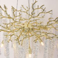 A close-up view of an ornate chandelier featuring gold-toned metal branches and cascading crystal elements. The chandelier's structure mimics organic tree branches with smooth, polished gold-colored metal forming an intricate network of curved and forked shapes. Hanging from these golden branches are multiple strands of clear, faceted crystals arranged in cylindrical formations. Each crystal strand resembles a delicate icicle or a string of diamonds, creating a shimmering, waterfall-like effect. Small, warm-toned LED light sources are visible nestled within the structure, illuminating the piece from within and enhancing the play of light on the crystal elements. The central mounting point of the chandelier is visible, showing how the golden branches radiate outward. The combination of the organic gold forms and the geometric crystal arrangements results in a striking blend of natural and luxurious design elements, suitable for high-end, contemporary interiors.