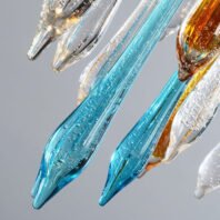 A close-up view of exquisitely crafted glass elements resembling icicles or elongated teardrops. The image features several glass pieces in various colors, including vibrant turquoise blue, amber, and clear. The glass has a textured appearance with tiny bubbles trapped inside, giving it a sparkling, crystalline quality. The shapes are elongated and tapered, ending in sharp points. Light reflecting off and through the glass creates a luminous effect, highlighting the craftsmanship and delicate nature of these chandelier components. The background is a soft, neutral gray, allowing the colorful glass pieces to stand out prominently.