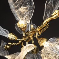 Detailed view of a chandelier featuring intricate gold-plated branches with delicate glass leaf-shaped shades. The transparent glass leaves have a veined texture, resembling real foliage. Gold fixtures hold small light bulbs, creating a warm glow. The design merges organic forms with luxurious materials, showcasing a blend of nature-inspired artistry and modern lighting craftsmanship against a dark background.