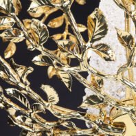 A close-up view of an opulent chandelier featuring intricate gold-plated metalwork in the shape of leaves and branches. The image showcases highly polished gold-toned metal formed into delicate, lifelike leaves with visible veins and natural-looking curves. These golden leaves are arranged in clusters along twisting branches, creating a lush, organic appearance. The metalwork has a brilliant, mirror-like finish that reflects light, emphasizing the sculptural quality of each leaf. Interspersed among the golden foliage are glimpses of sparkling crystal elements. These crystals appear to be cut in irregular, rock-like shapes, providing a striking contrast to the smooth, polished gold. The crystals catch and refract light, adding a dazzling dimension to the overall design. In the background, parts of what seems to be the chandelier's main structure are visible, including what looks like a gold-plated mounting bracket or central stem. The dark background in parts of the image helps to accentuate the brilliant gold tones and the sparkle of the crystals. This chandelier combines the warmth and luxury of gold with the brilliance of crystal, resulting in a piece that is both nature-inspired and opulent. It would serve as a stunning focal point in high-end interior spaces, bringing a touch of natural elegance with its leaf motif while exuding luxury through its materials and craftsmanship.