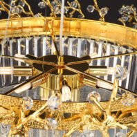 A close-up view of an opulent chandelier combining gold-plated elements with crystal accents. The central focus is a modern starburst light fixture with six golden arms radiating outward, each holding a cylindrical light bulb. This central piece is surrounded by a circular gold-plated frame with intricate, textured detailing. Hanging from this frame are numerous rectangular crystal prisms, creating a shimmering curtain effect. Above the main frame, golden branches with a organic, vine-like design extend outward, adorned with small crystal orbs. The chandelier's structure showcases a harmonious blend of contemporary and classic design elements, with the sleek, modern central light fixture contrasting beautifully against the more traditional gold filigree and crystal elements. The gold plating has a rich, warm tone that complements the brilliance of the crystal components. This chandelier would serve as a stunning focal point in any luxury interior, combining the timeless elegance of crystal with the warmth of gold and a touch of modern design.