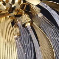 A close-up view of an ornate chandelier showcasing a blend of gold and crystal elements. The image captures the intricate details of the fixture, highlighting its luxurious design. Gold-toned metal parts with ribbed textures form petal-like shapes, contrasting beautifully with clear crystal components that have delicate, vein-like patterns. Small, bell-shaped golden casings house LED lights, creating a warm glow. Crystal 'stems' extend from these casings, adding to the floral motif. The chandelier's elements overlap and intertwine, creating a complex, layered effect that mimics the organic structure of a flower. The combination of polished gold surfaces and transparent crystal creates a play of light and shadow, emphasizing the chandelier's three-dimensional quality. This detailed view demonstrates the meticulous craftsmanship involved in creating this high-end, nature-inspired lighting fixture, which seamlessly blends modern luxury with organic forms.