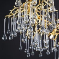 An exquisite branch chandelier featuring a golden, tree-like structure adorned with numerous crystal pendants. The chandelier's frame is crafted from intricately designed gold-toned metal, resembling delicate tree branches. Suspended from these golden branches are an array of clear crystal pendants in various lengths and shapes. The pendants feature long, slender stems ending in bulbous drops, reminiscent of raindrops or dewdrops. These crystal elements catch and refract light, creating a dazzling display against the warm golden structure. The image captures the chandelier's lower portion, showcasing the interplay between the solid, gleaming gold branches and the transparent, sparkling crystal droplets. This branch chandelier seamlessly blends organic forms with luxurious materials, offering a stunning focal point that brings the beauty of nature indoors in an opulent, contemporary style.