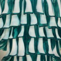 A close-up of a large chandelier featuring custom lighting design. Alternating layers of teal green and white glass elements create a mesmerizing pattern. The hotel chandelier's unique composition resembles cascading waves or overlapping leaves. Each piece of this big chandelier has a curved, elongated shape with smooth edges, showcasing exquisite craftsmanship in custom lighting for upscale hospitality spaces.