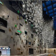 A breathtaking large chandelier dominates a unique hotel interior, showcasing innovative custom lighting design. This big chandelier features hundreds of suspended crystal cubes, creating an illusion of frozen raindrops or icicles. The chandelier stretches along the ceiling, its transparent elements catching and refracting light brilliantly. Below, a striking concrete wall is adorned with an array of colorful, irregularly placed windows and recesses, some illuminated in vibrant hues. The juxtaposition of the ethereal chandelier against the industrial-chic wall creates a captivating contrast, embodying contemporary luxury and artistic vision in hotel design.